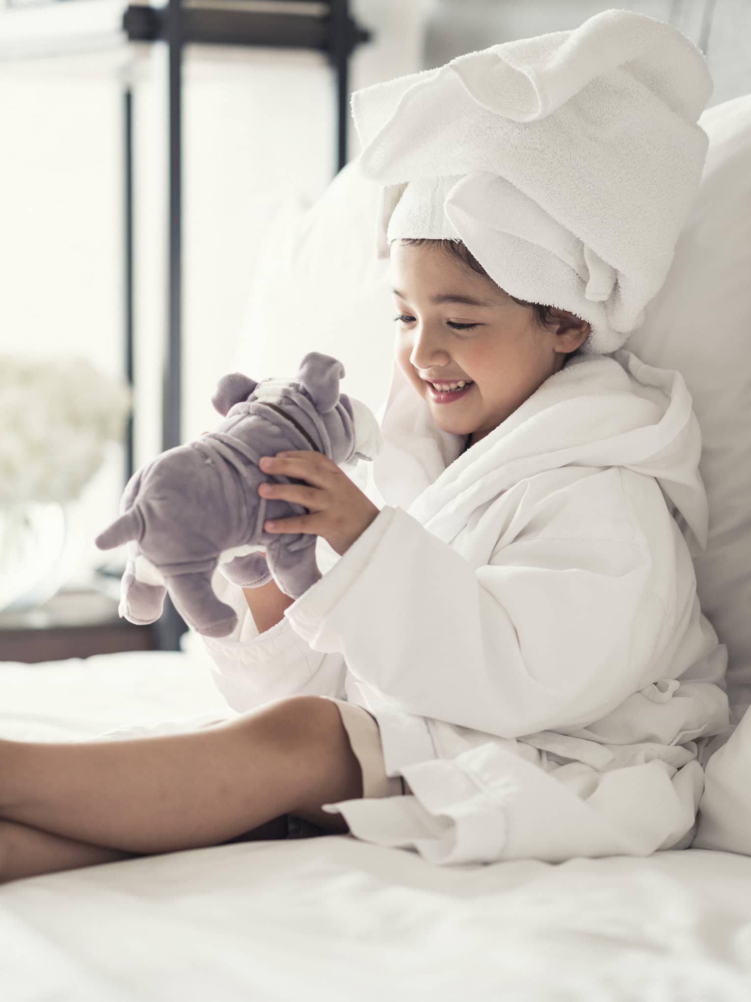 Young girl wearing a white bathrobe with towel wrap plays with plush dog toy on bed