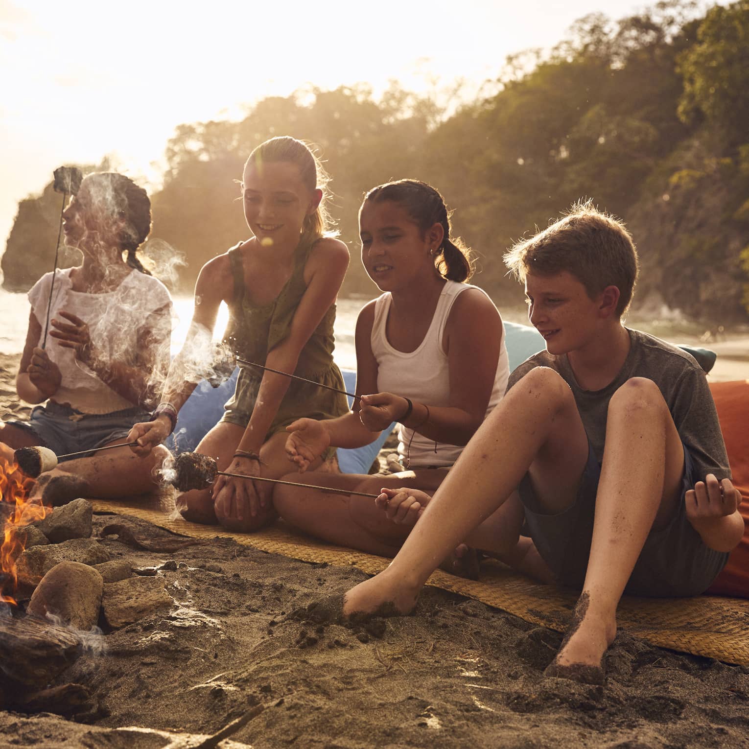 A group of young kids on a beach sitting near a small fire.