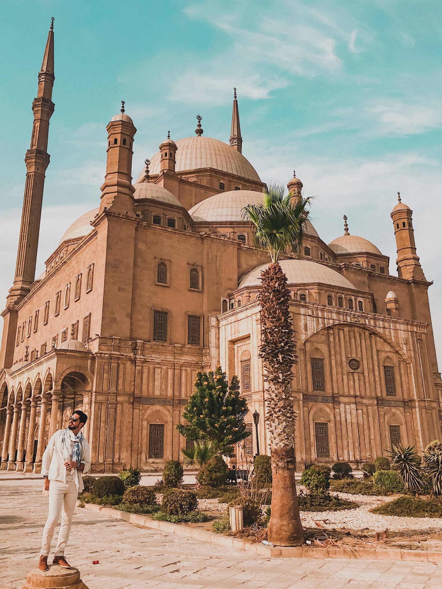 A tourist poses in front of the imposing Mosque of Muhammad Ali, minarets towering above the many domes of the building.