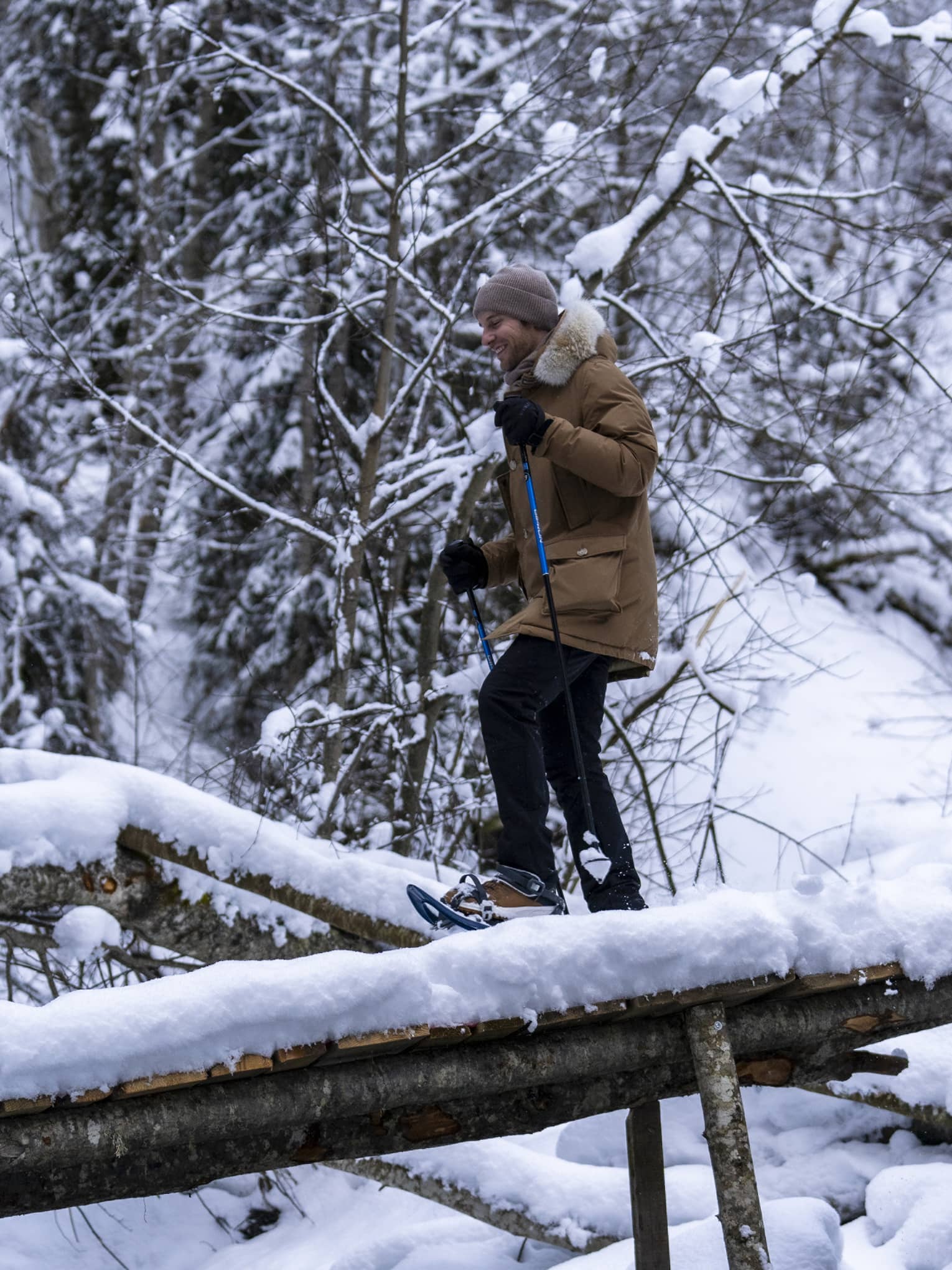 A snowshoer in a knit cap, parka and gloves uses poles to cross fallen logs in a dense, snow-covered forest.