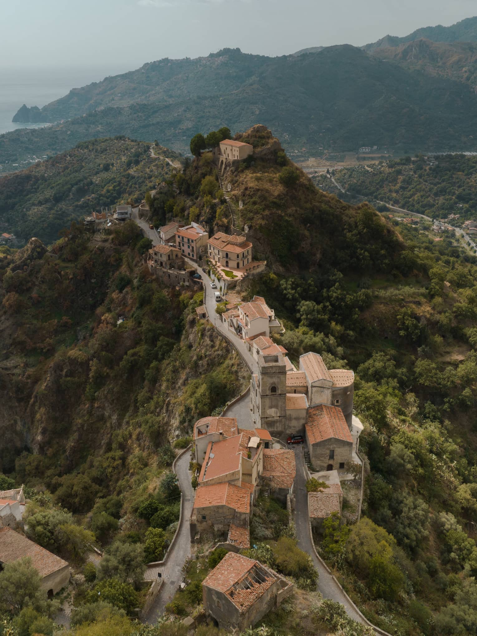 Aerial view of a historic hilltop village with winding roads, stone buildings and terracotta roofs, nestled in a lush green mountain landscape.