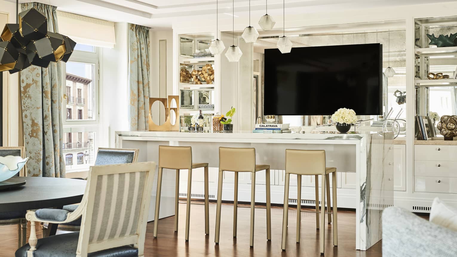 Luxury hotel suite lounge area with a marble counter, beige barstools, mounted TV, decorative shelving, dining table and large windows with patterned curtains