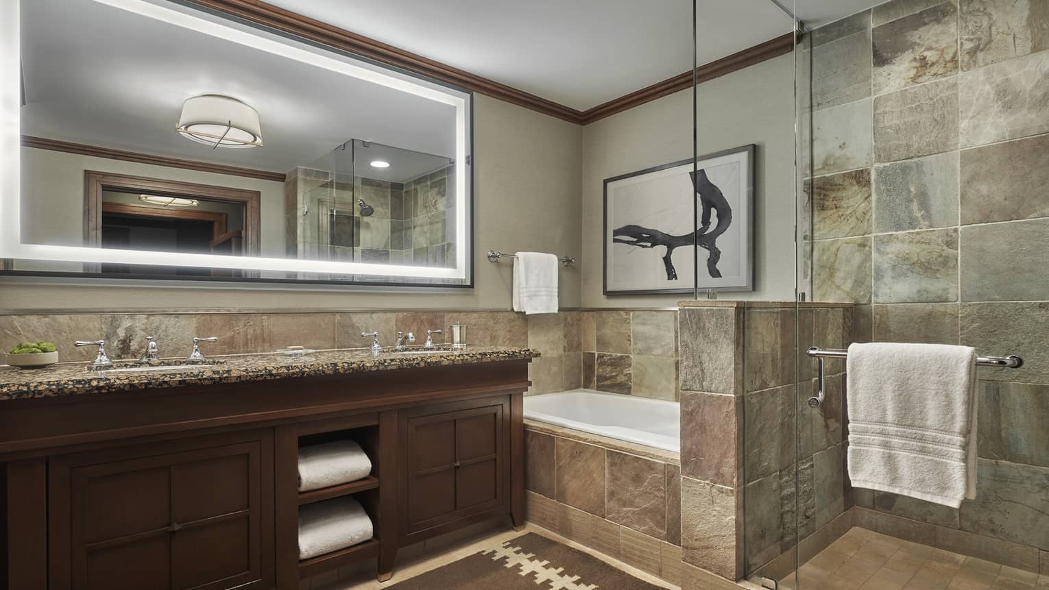 A tiled bathroom, with a double vanity, large lighted mirror and a shower and tub separated by a glass partition