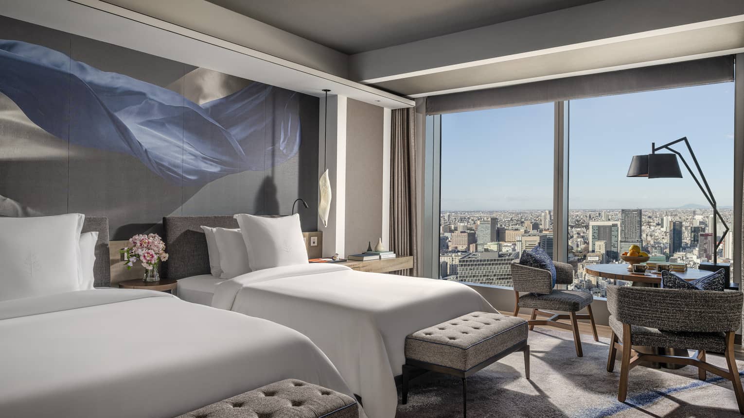 Guest room with two beds, upholstered benches and floor-to-ceiling windows offering panoramic city view, featuring modern décor