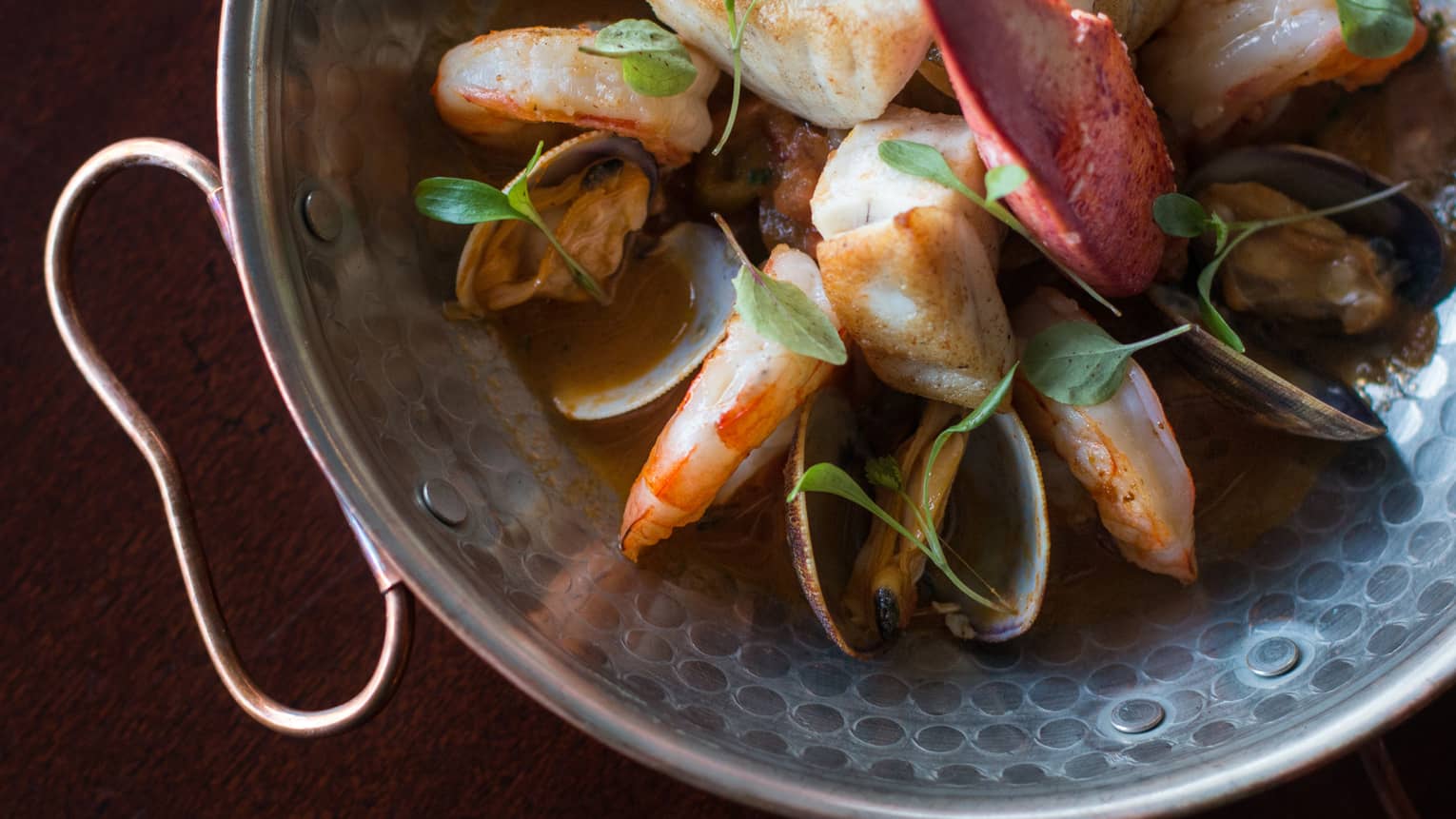 Close-up of an elegant copper handle on a patterned metal bowl of seafood in a rich, dark broth garnished with green herbs.