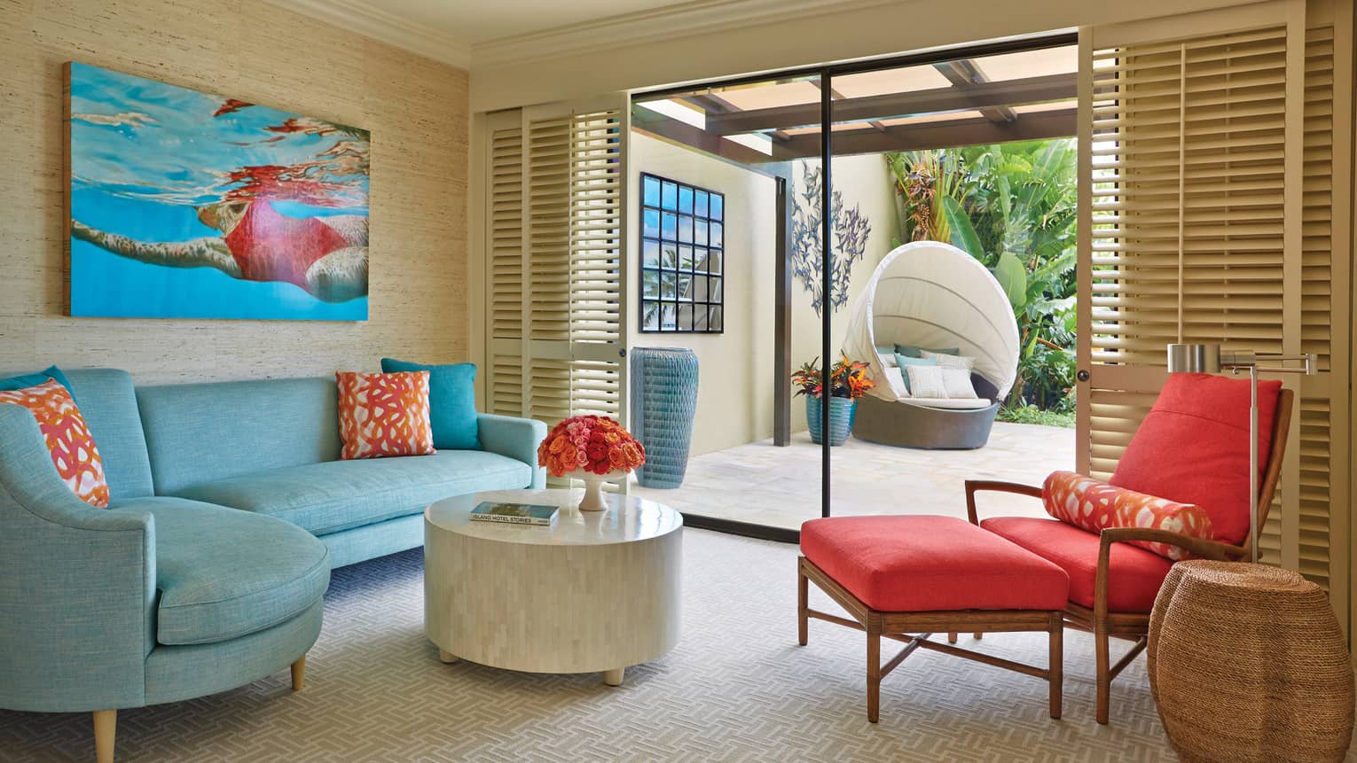 Seating area with a blue sofa, colorful pillows and red chaise lounge, opening to a patio with a hanging chair and lush garden view at a luxury resort suite