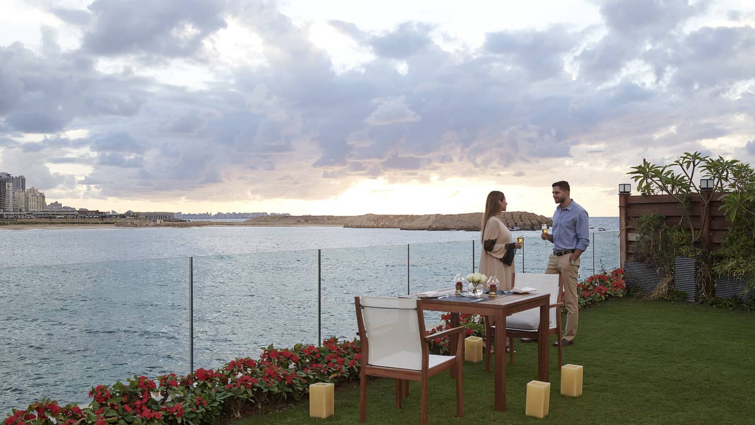 A standing couple chats at the water's edge at sunset, a table set for two in the foreground 