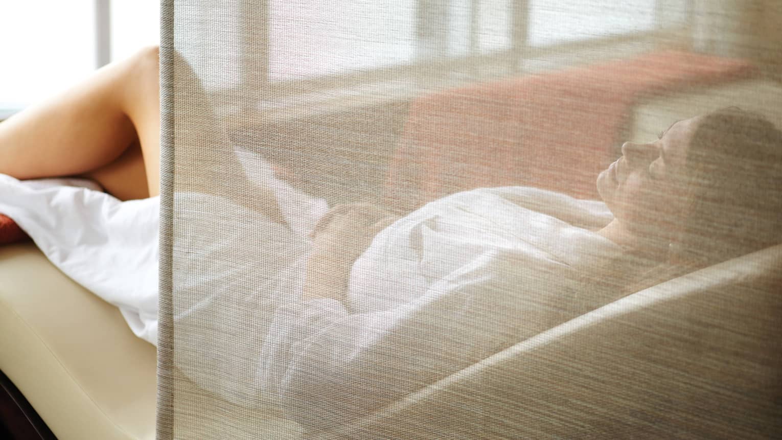Woman wearing white bathrobe lies on curved spa chair behind sheer curtain