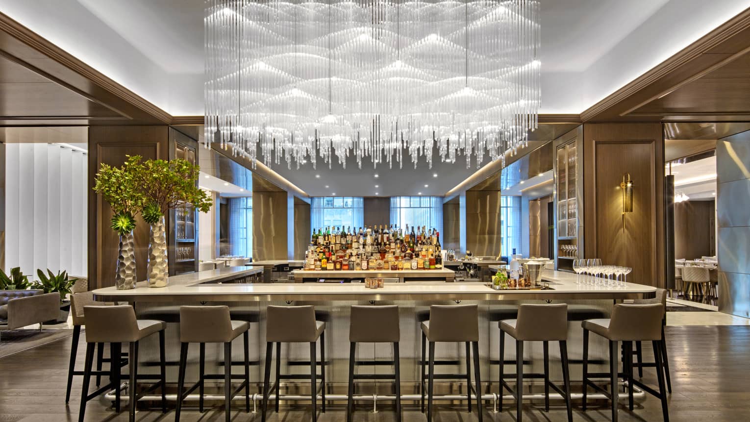 A large bar area with hanging glass chandelier and many grey stools.