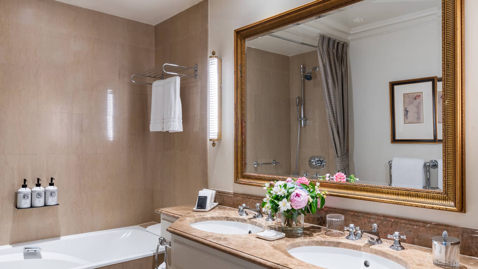 Luxury hotel bathroom with a marble countertop vanity, double sinks, fresh flowers, large framed mirror and bathtub with wall-mounted toiletries 