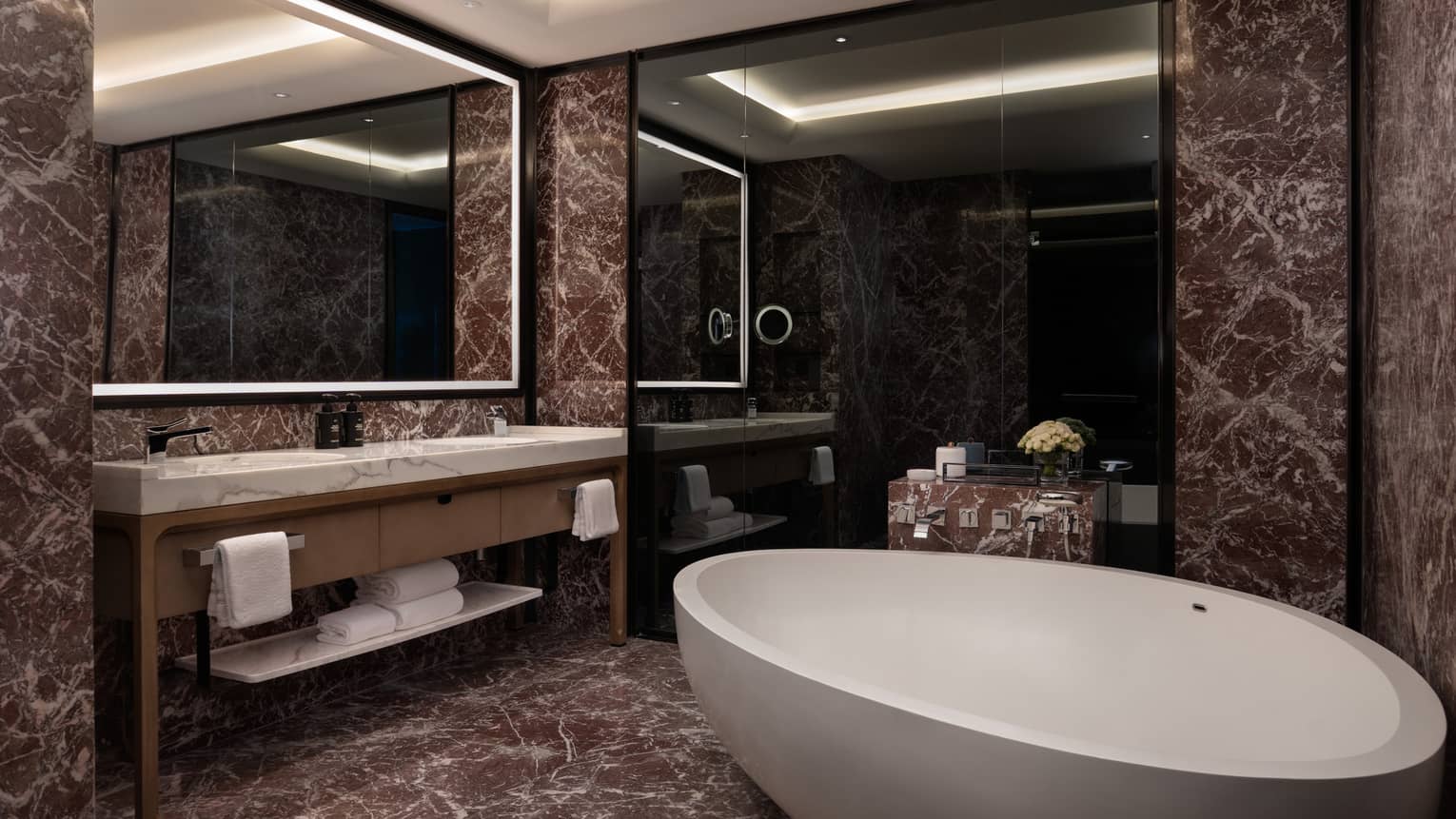 Bathroom with a large standalone tub, dark marble tiles and an illuminated mirror over double vanity