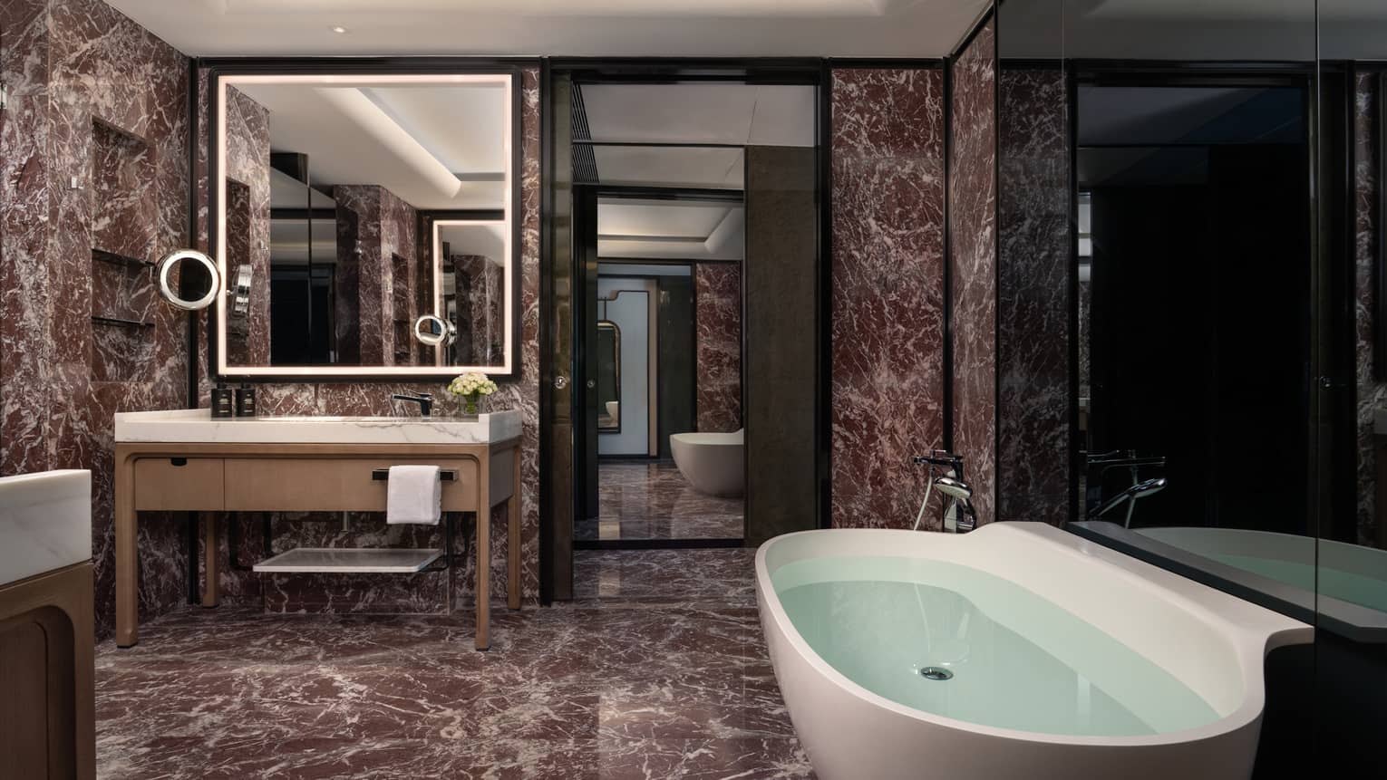 Sleek bathroom featuring dark marble tiles, a standalone tub and an illuminated square mirror over the vanity