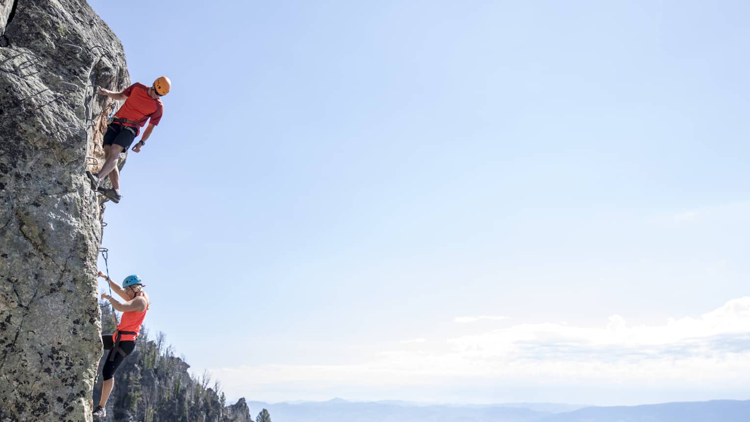 Man and woman climb steep cliff face with view of mountains in far distance 