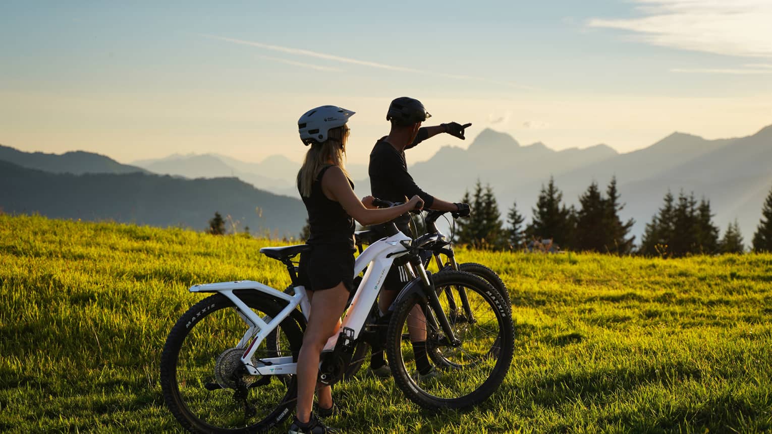 Two people riding fat bikes on meadow, one pointing to distant mountains