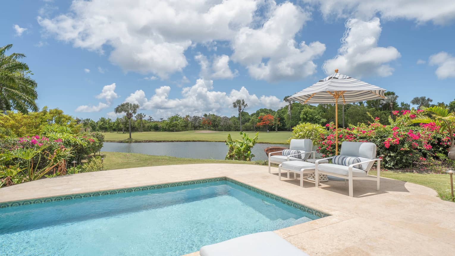 Pool deck with a rectangular pool, two arm chairs under striped umbrella, pond in the distance