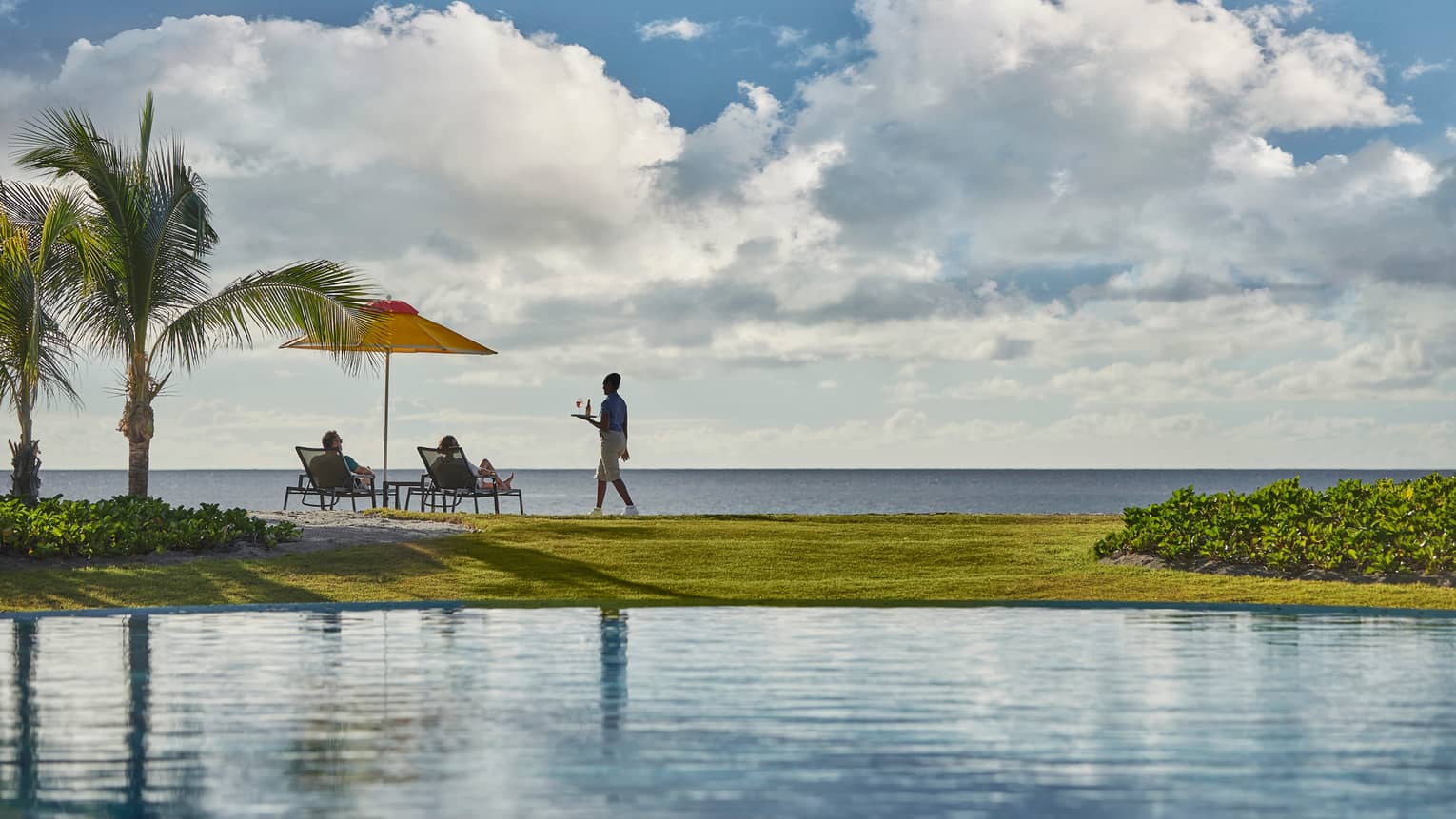 View across pool to green lawn, waitstaff carrying tray to couple in patio chairs overlooking ocean