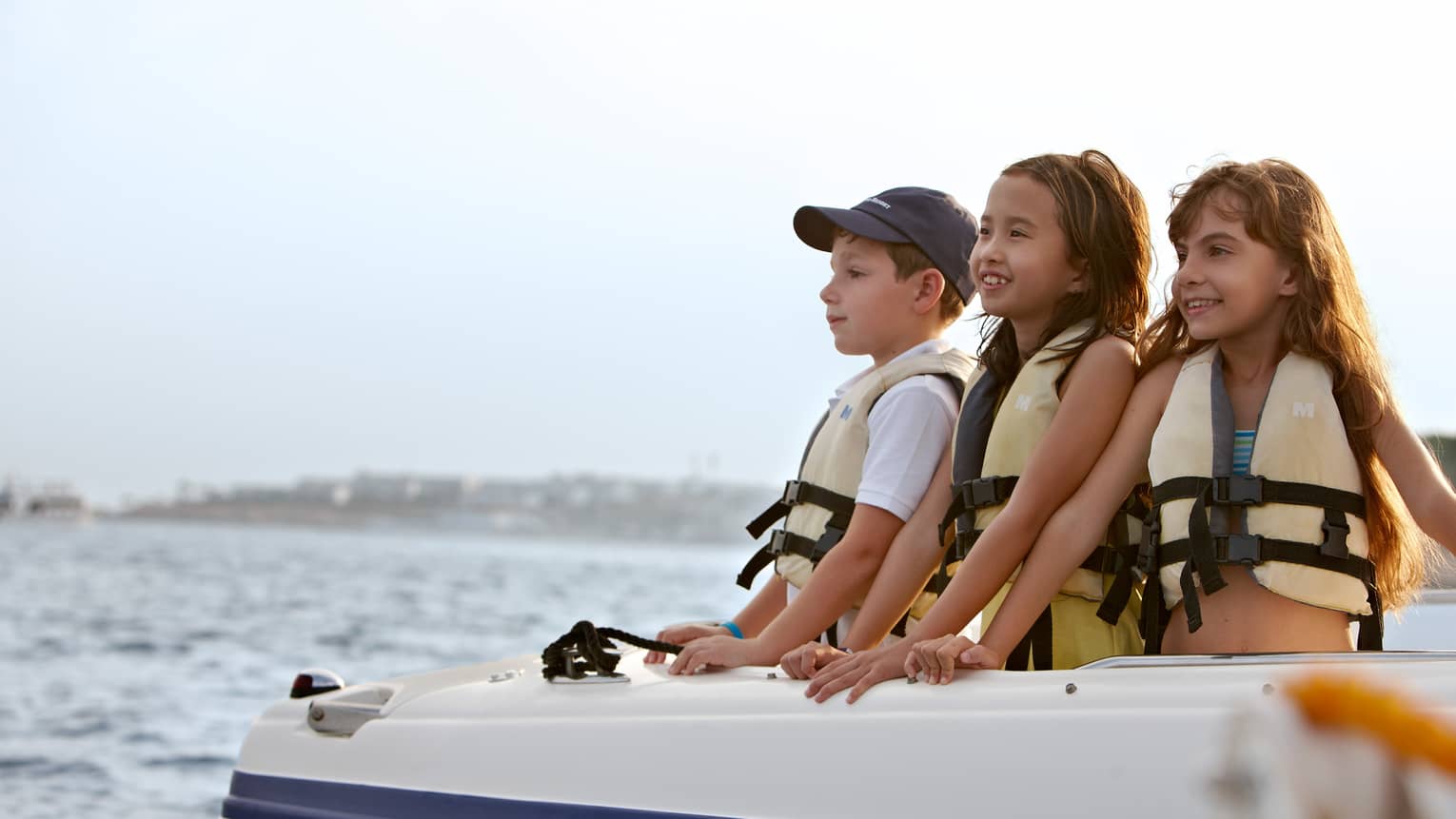 Three children in lifejackets in front of boat smile, look out on water 