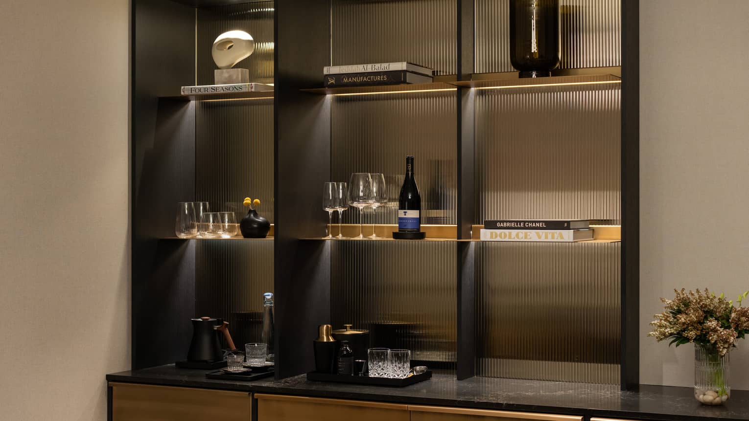 Hotel suite bar shelf with books, glasses, wine bottle, decorative pieces and a flower vase