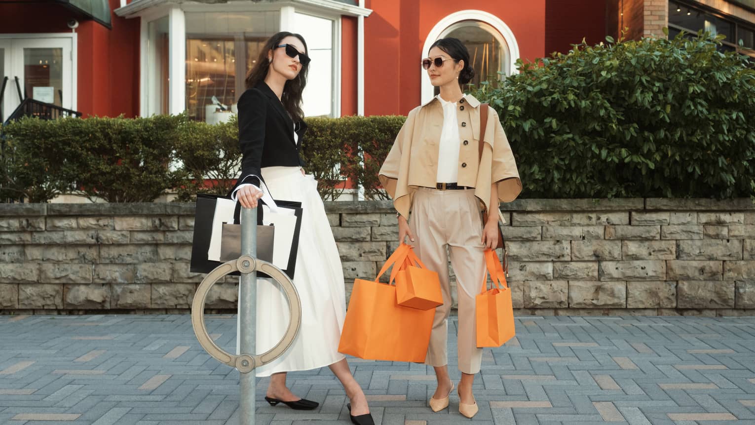 Two stylish people with shopping bags, standing outdoors in front of a red building, dressed in chic, neutral-coloured outfits and sunglasses