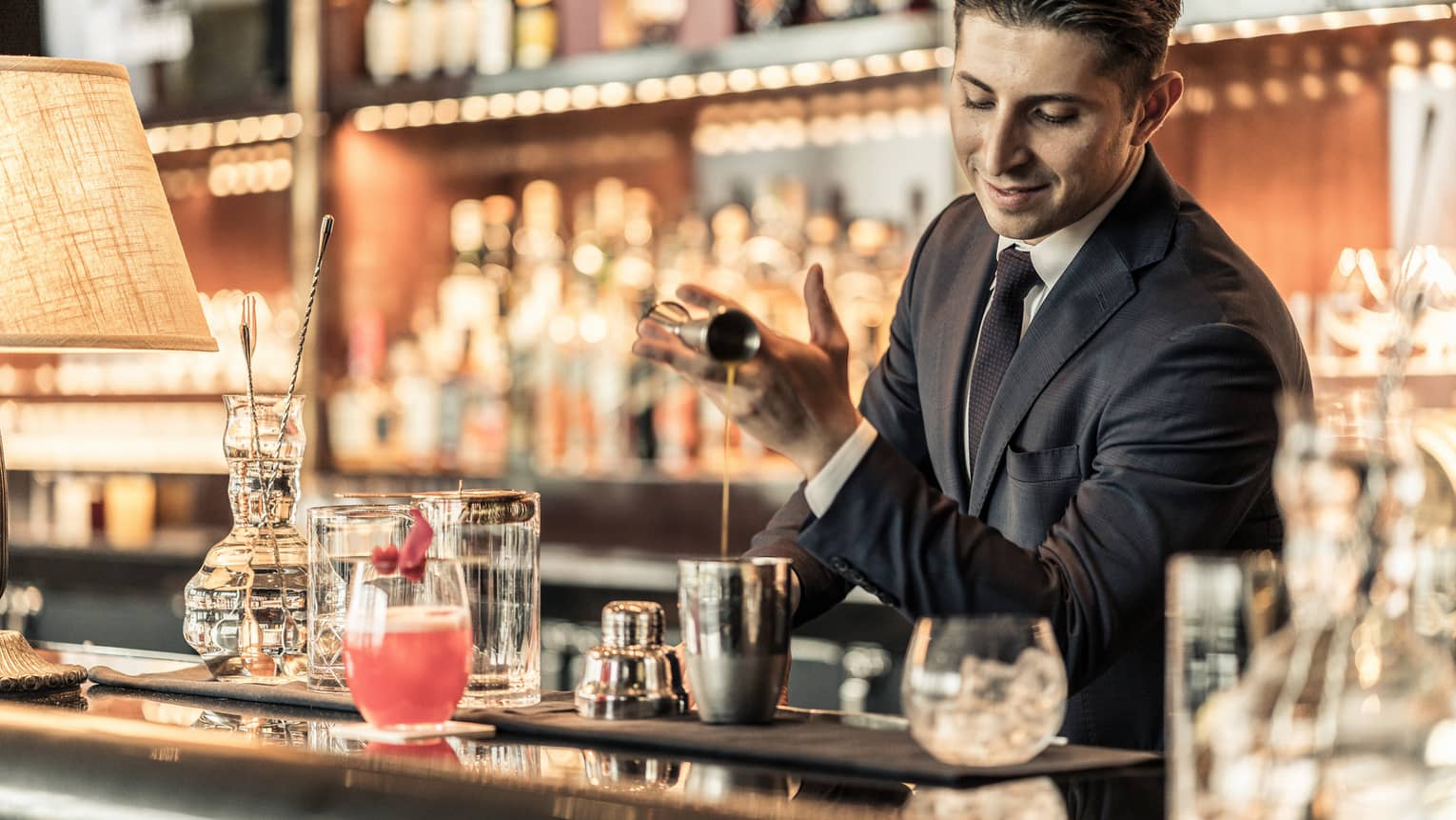 Bartender behind bar, pouring liquid from jigger into metal shaker among bar accessories