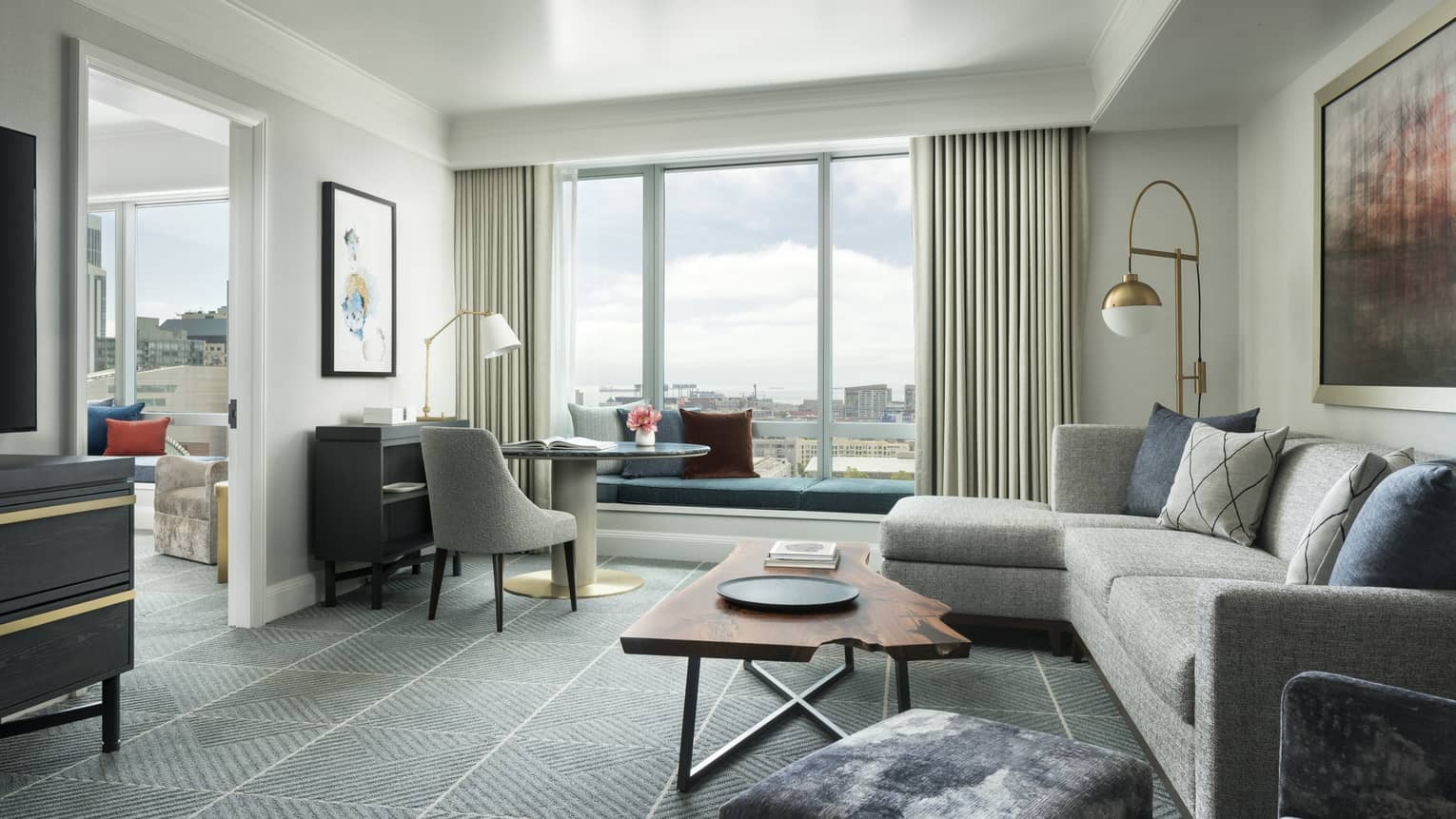 Hotel suite separate living room with retro-style L-shaped grey sofa and desk chair, live-edge wood coffee table