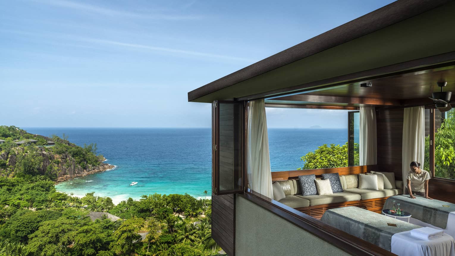 Woman smoothes sheet on side-by-side massage tables in spa room high above trees, ocean