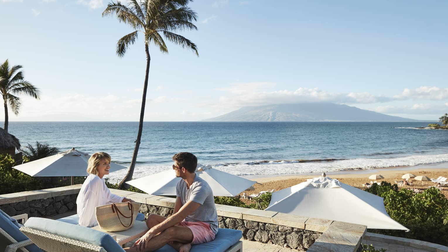 Man in shorts and smiling woman in beach shirt sit on edge of lounge chair, beach and ocean in background