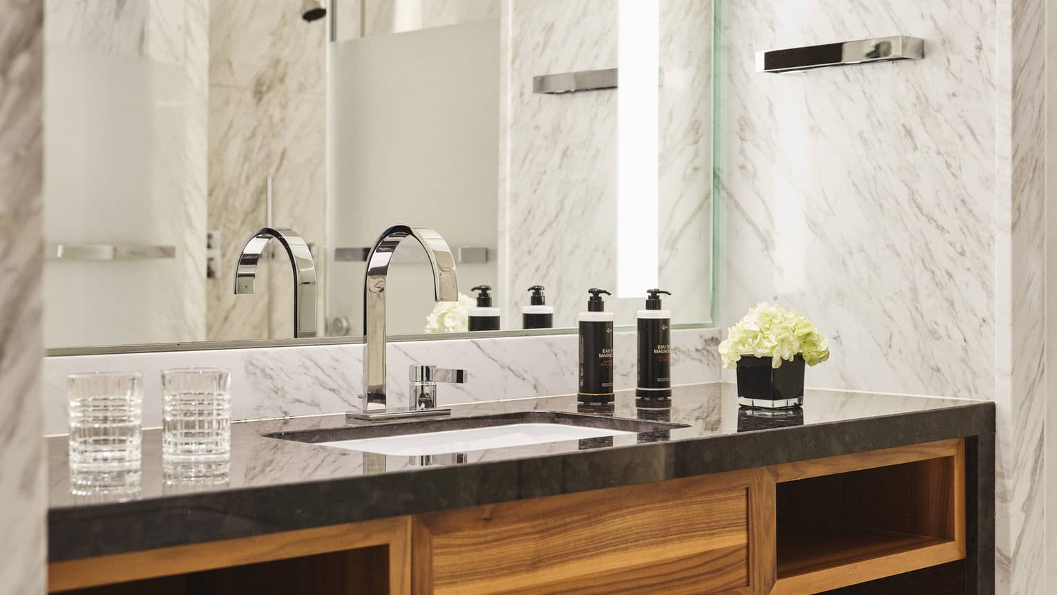 Hotel bathroom vanity with black countertop, chrome faucet, wooden shelving, two glasses, toiletries, a small bouquet of white flowers, mirror and marble walls