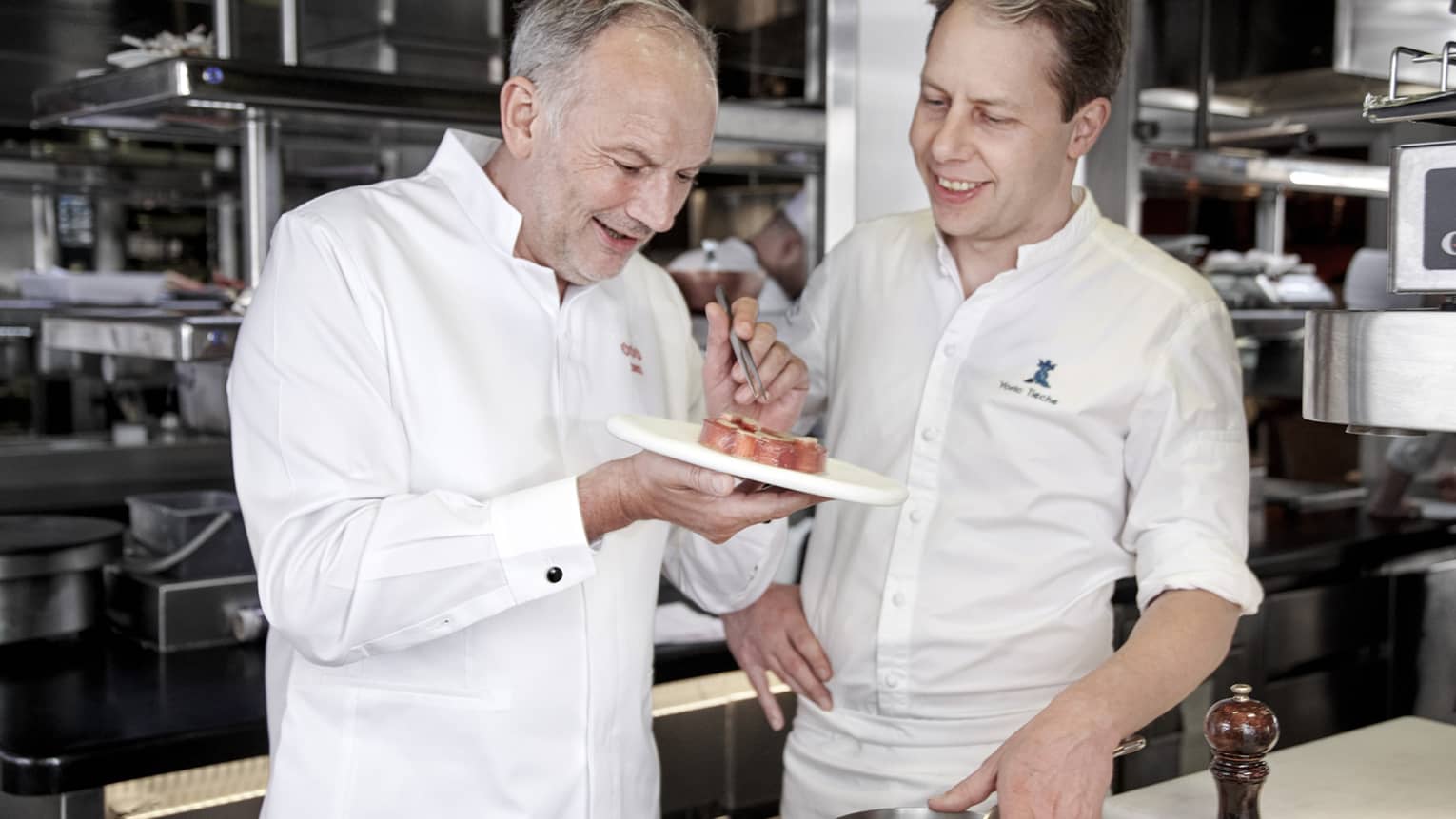 Two smiling chefs garnish small dish with tweezers in kitchen