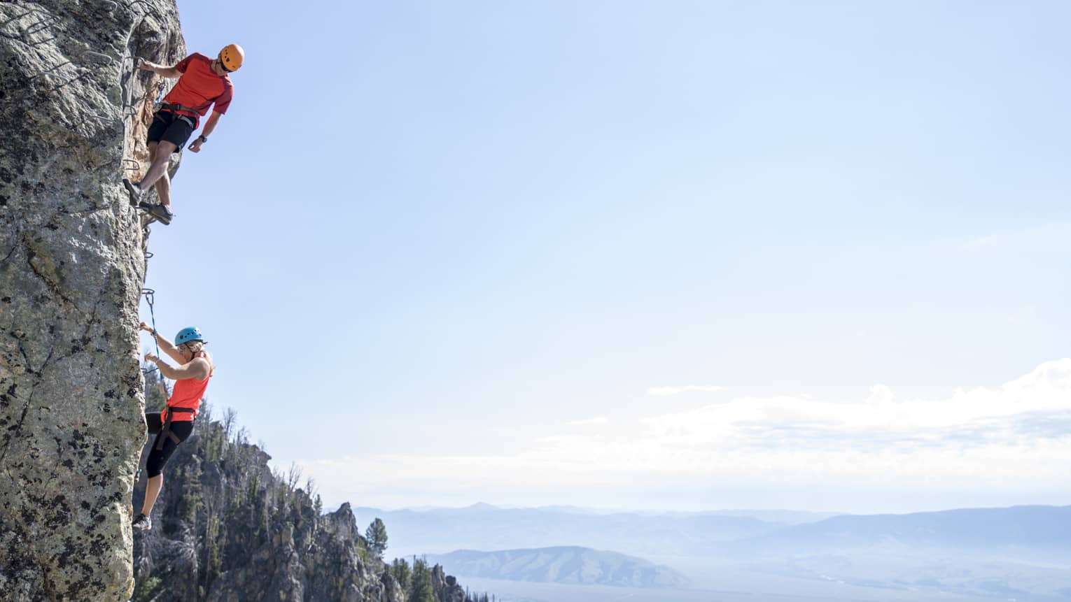 Man and woman climb steep cliff face with view of mountains in far distance 