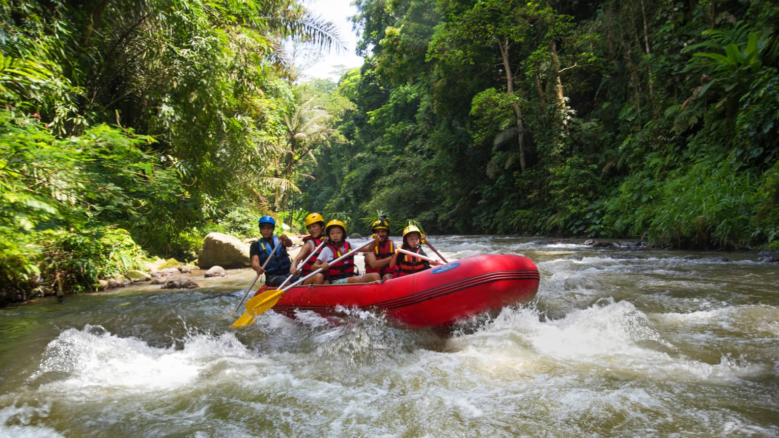 Family of four and guide hold paddles, steer red inflatable raft on rapids in river