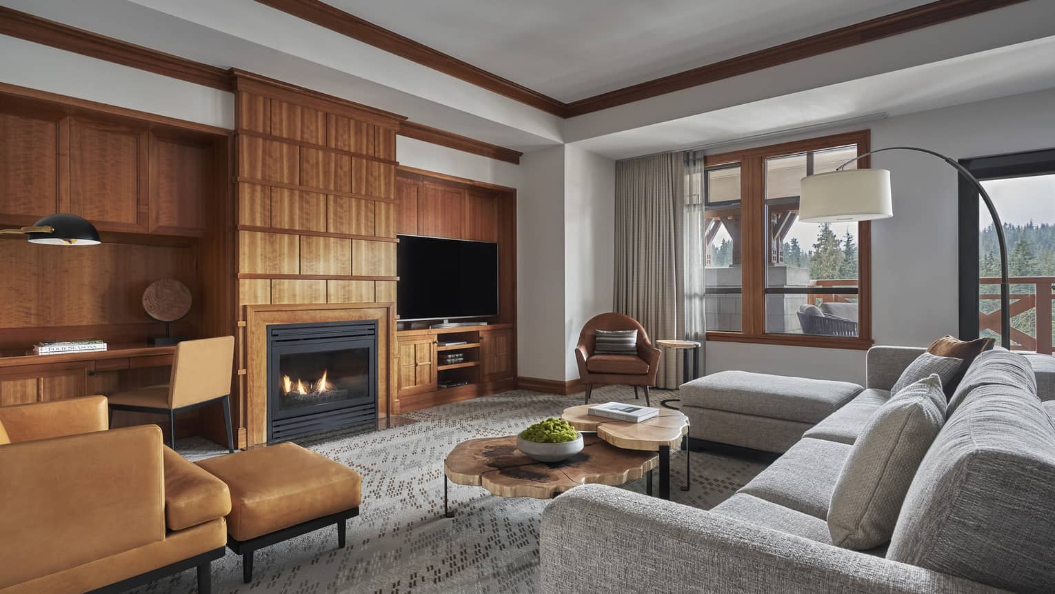 The Alpine Suite at Four Seasons Whistler featuring a sophisticated and cozy living area. The room is decorated with natural tones and includes a plush gray sectional sofa, a tan leather armchair with a matching ottoman, and wooden shelving units filled with books and decorative items. A contemporary fireplace and a large flat-screen TV mounted above it create a warm focal point. Large windows offer views of the mountainous landscape, contributing to the room’s tranquil ambiance.,The Alpine Suite at Four Seasons Whistler featuring a sophisticated and cozy living area. The room is decorated with natural tones and includes a plush gray sectional sofa, a tan leather armchair with a matching ottoman, and wooden shelving units filled with books and decorative items. A contemporary fireplace and a large flat-screen TV mounted above it create a warm focal point. Large windows offer views of the mountainous landscape, contributing to the room’s tranquil ambiance.