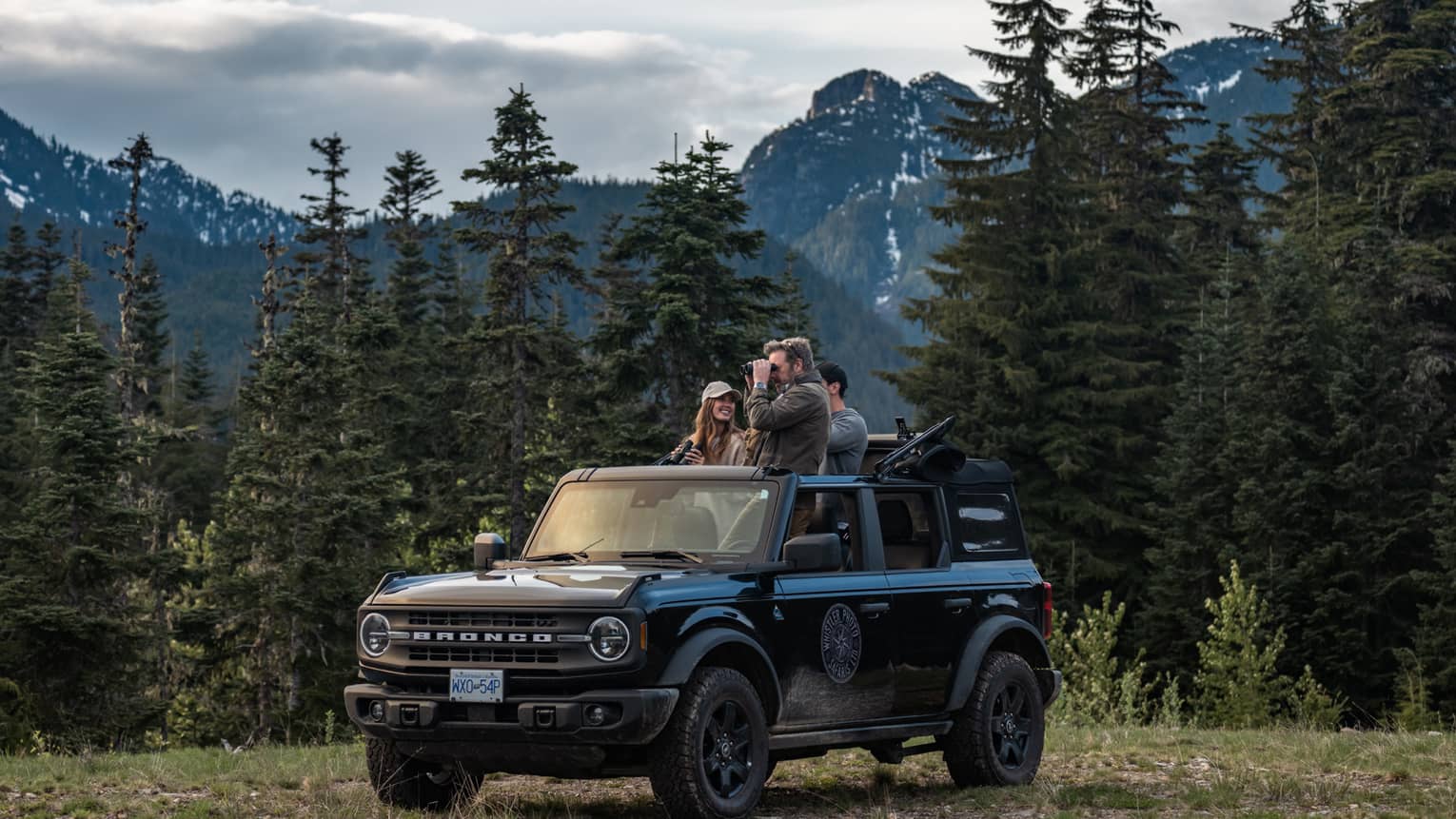Group of friends with binoculars stand in open-top Ford Bronco surrounded by trees and mountains