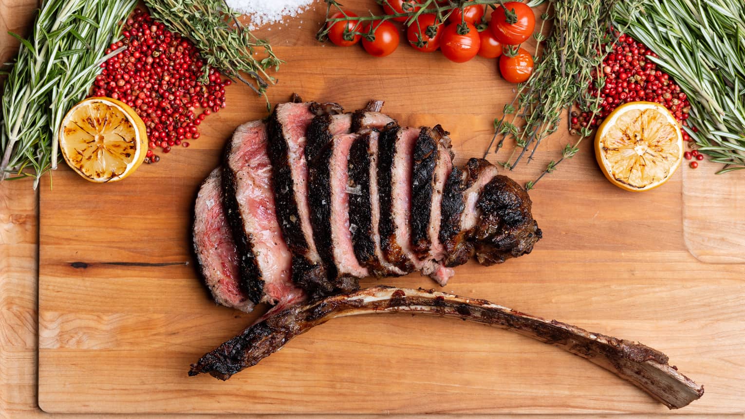 A wood serving platter holds slices of cooked meat along with garnishes including herbs, tomatoes and citrus.