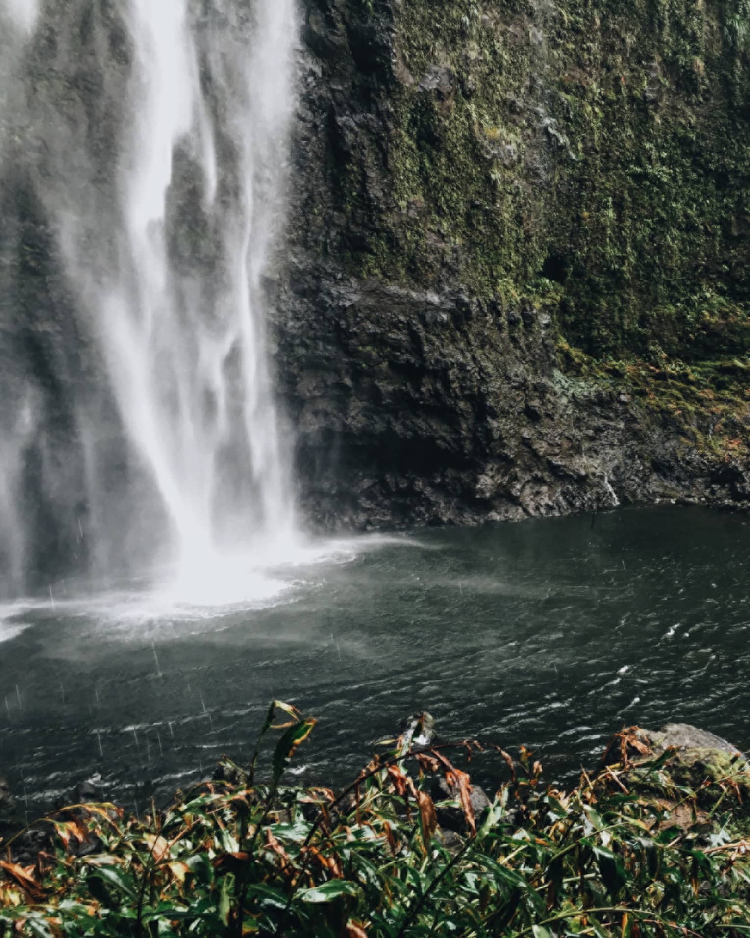 Back of woman hiking over black rocks under tall waterfall, small pond