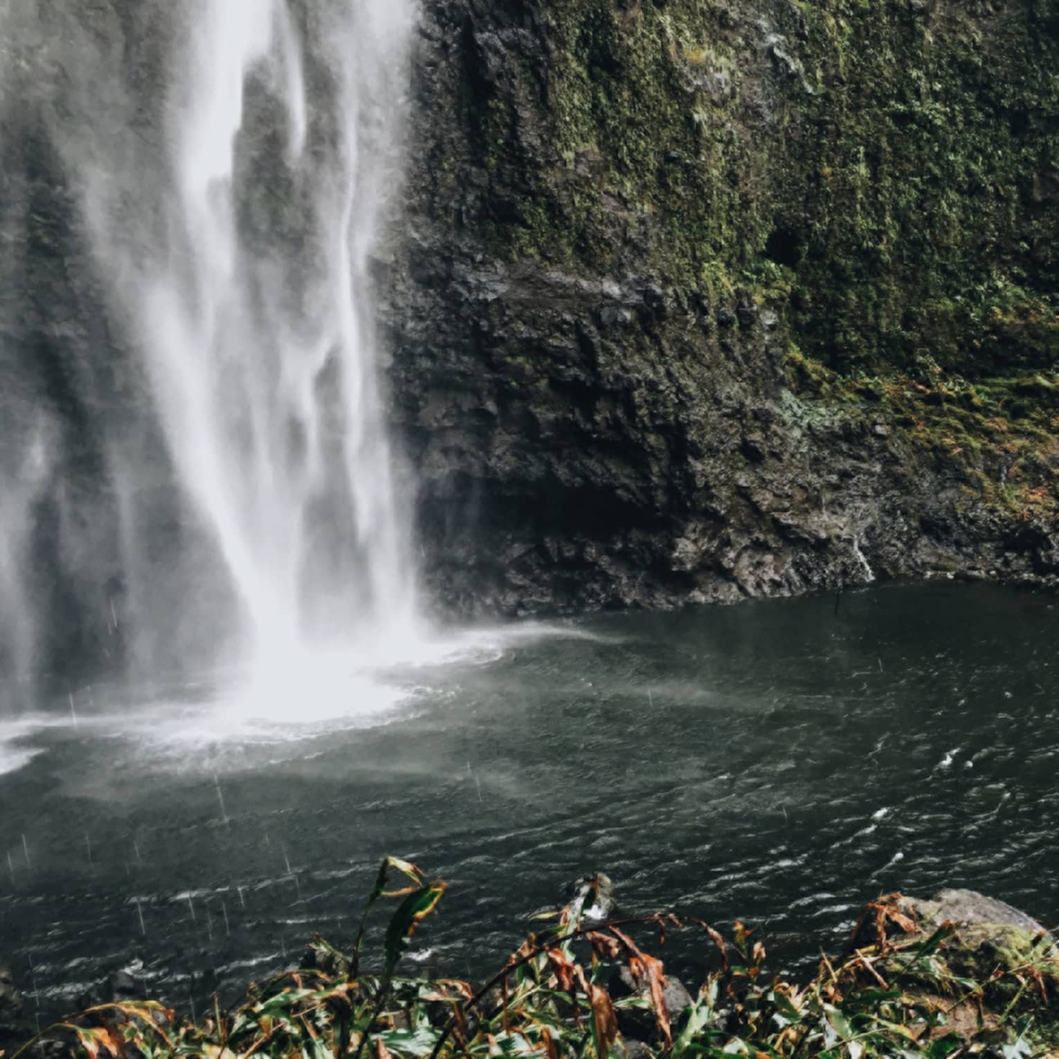 Back of woman hiking over black rocks under tall waterfall, small pond