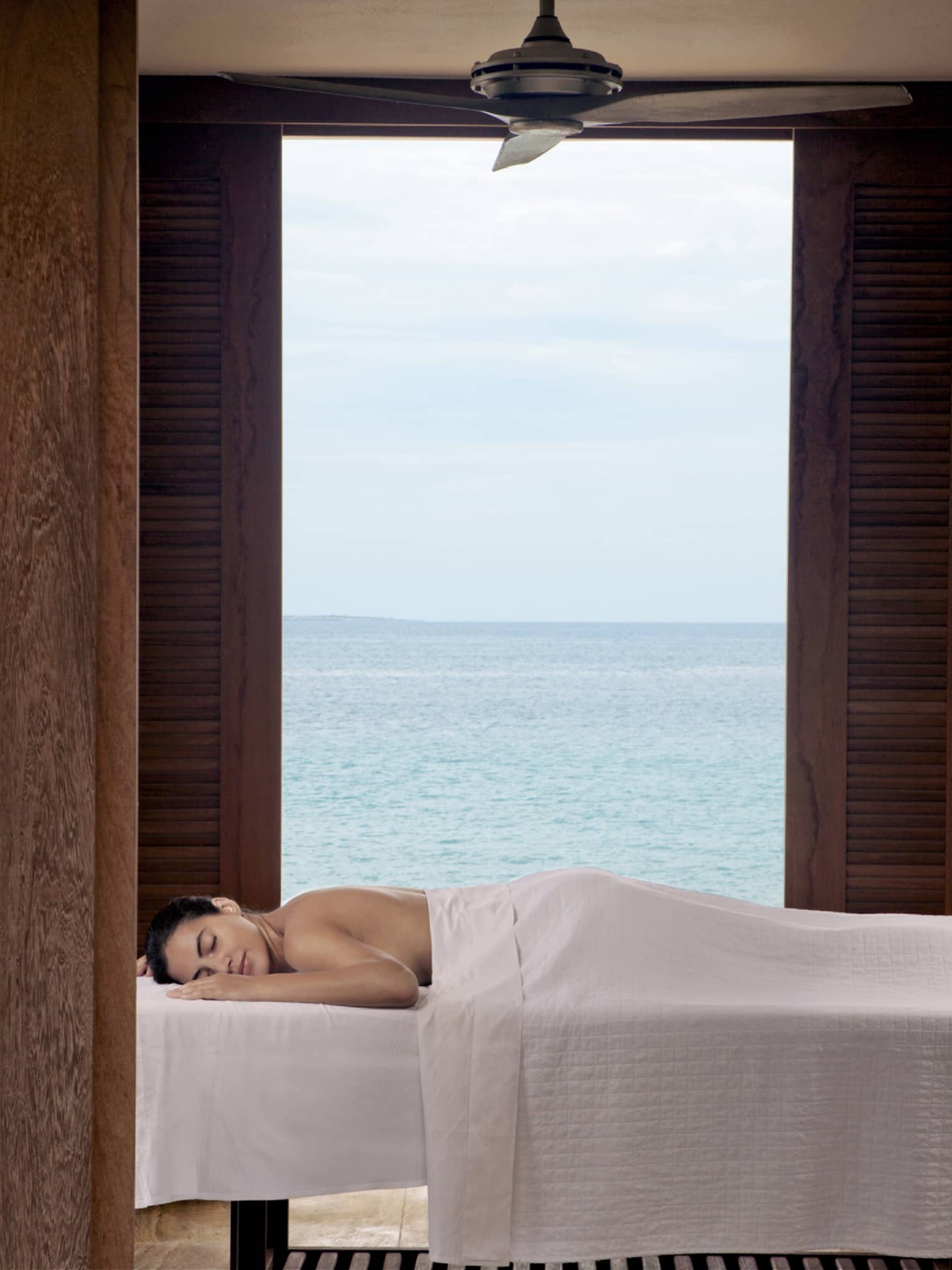 Guest enjoying a relaxing outdoor massage with a serene ocean view, framed by wooden shutters at a luxury beachfront resort