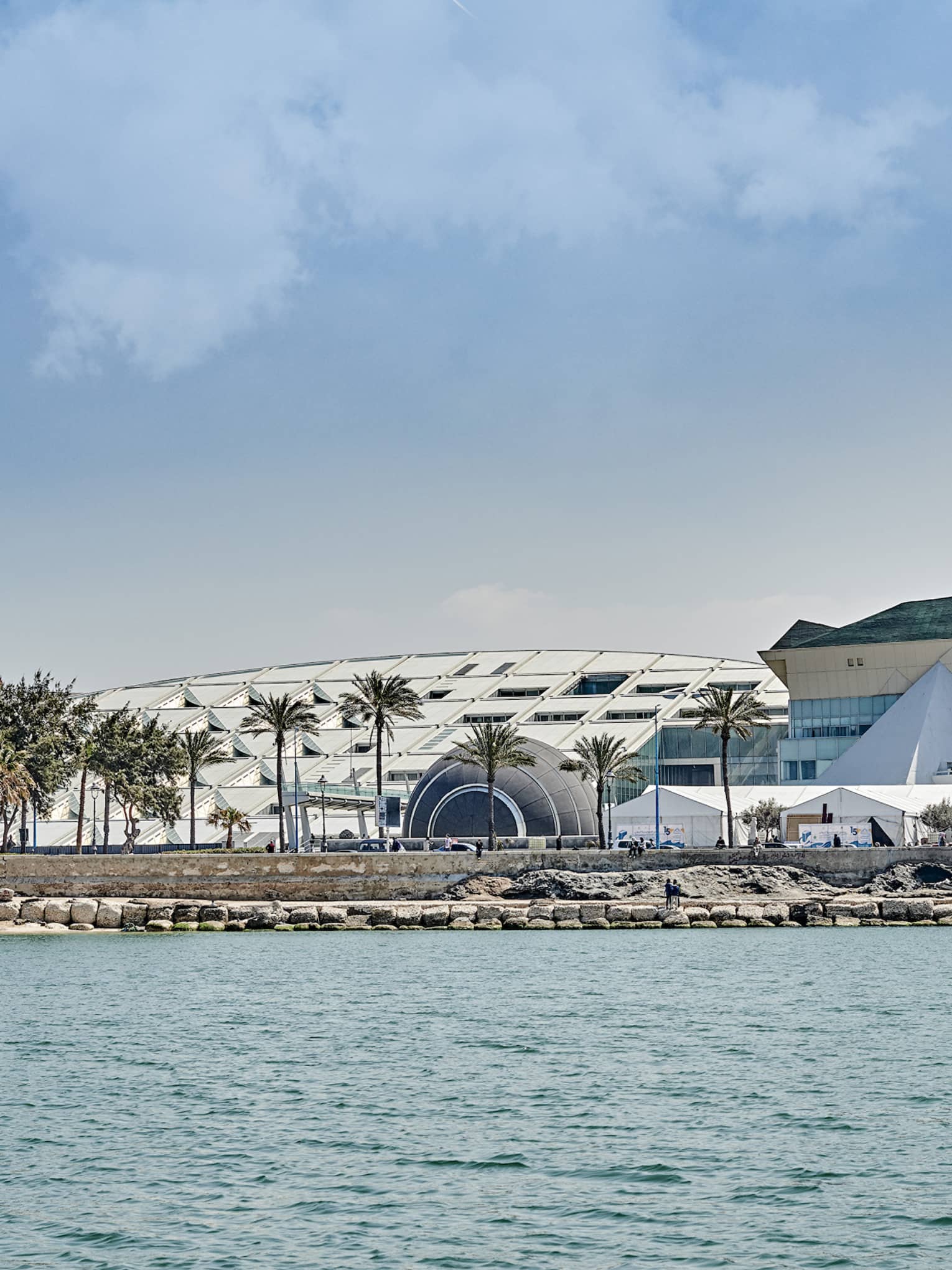 View over calm water towards a stony shoreline where people mingle under palm trees in front of a large, sloped building.