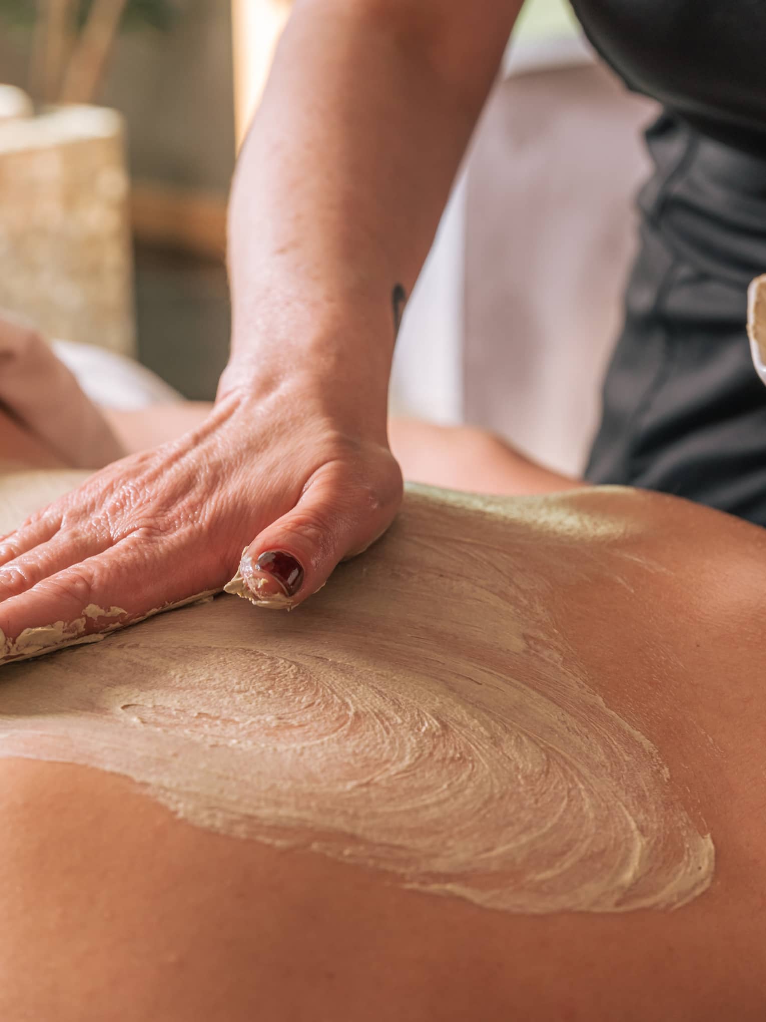 A hand rubs a sand-coloured body treatment on a person's bare back