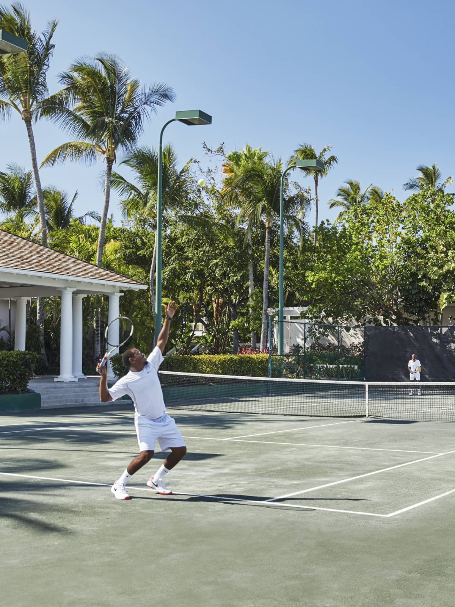 A man in all white trows a tennis ball high into the air, mid-serve