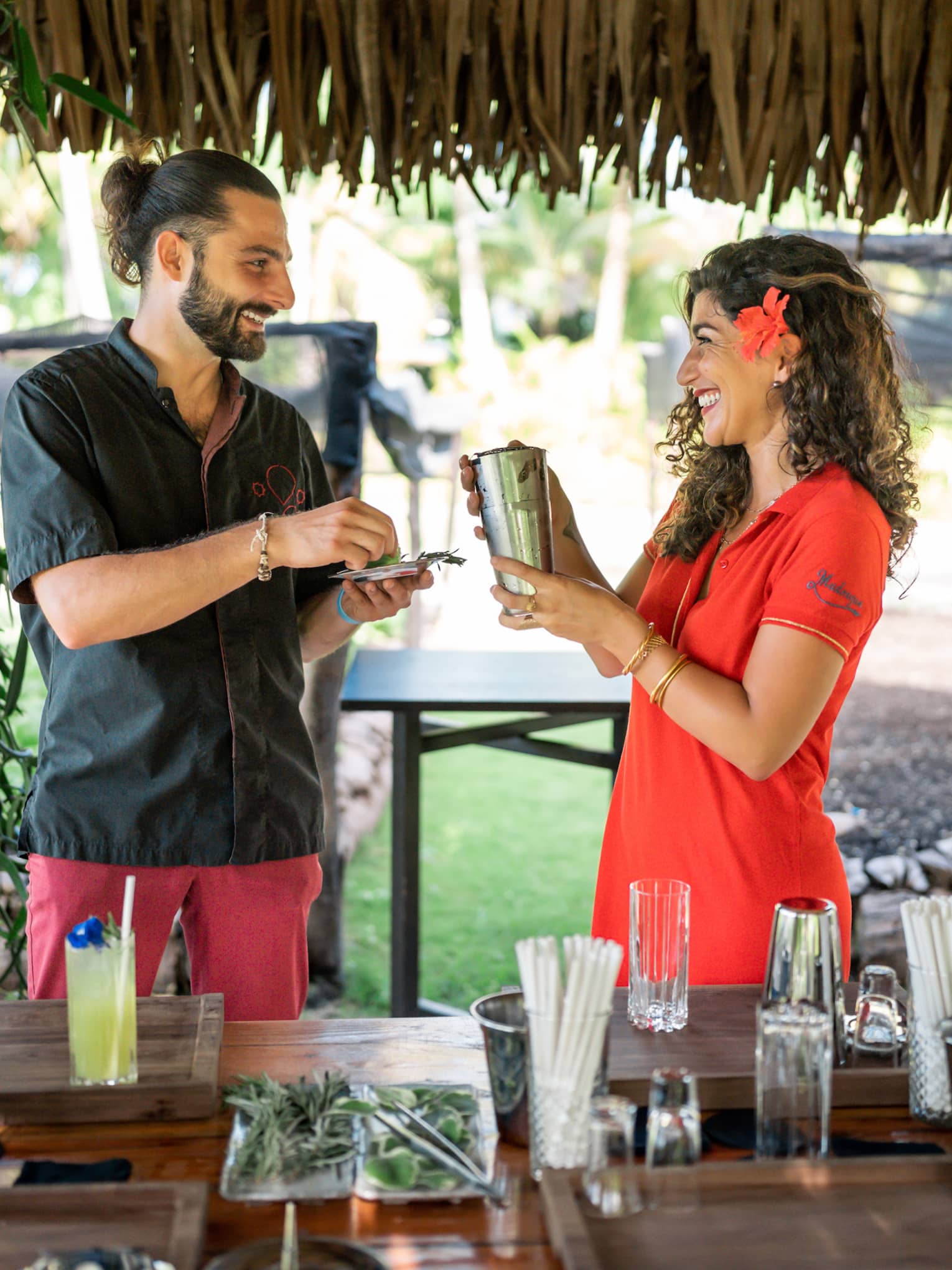 Two people hold a shaker and fresh herbs in a straw-roofed hut over a bar strewn with glasses, liquor bottles and garnishes.