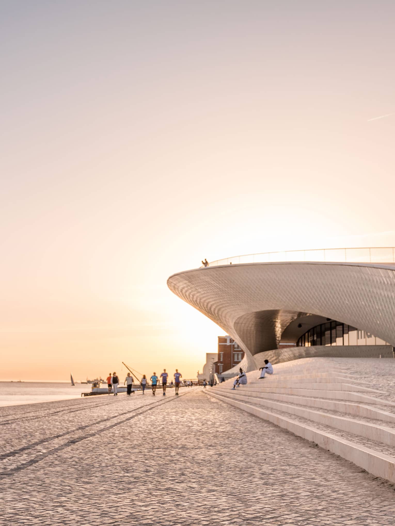 Modern waterfront building with sweeping architectural curves, people walking along a cobblestone promenade at sunset.