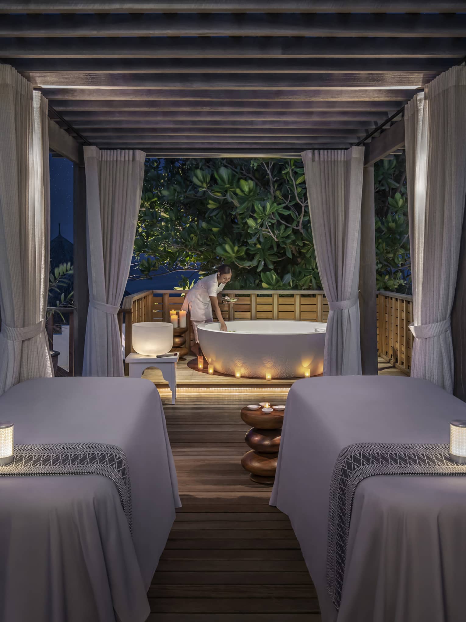 Outdoor night spa setting with two massage tables, soft candlelight, and a staff member preparing a bathtub under a canopy surrounded by tropical greenery