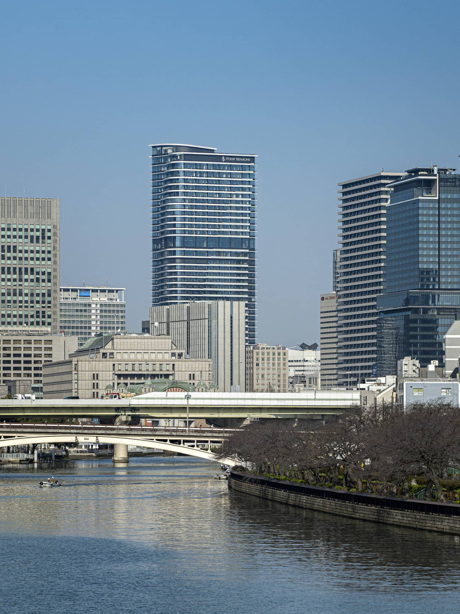 Under a clear blue sky, Osaka's modern high-rises loom above two bridges spanning a river lined with dormant cherry blossoms.