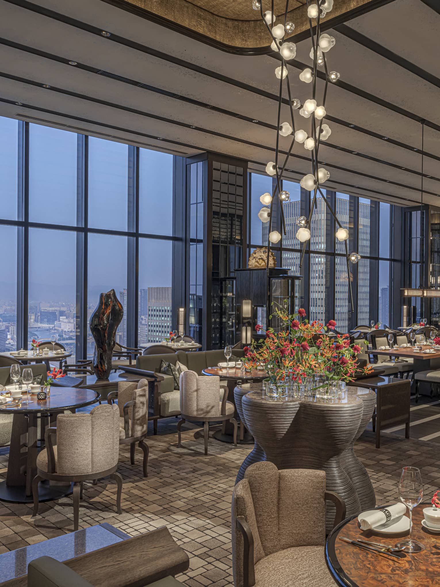 Expansive high-end restaurant warmly lit by modern chandeliers with a bank of windows offering aerial views of the city