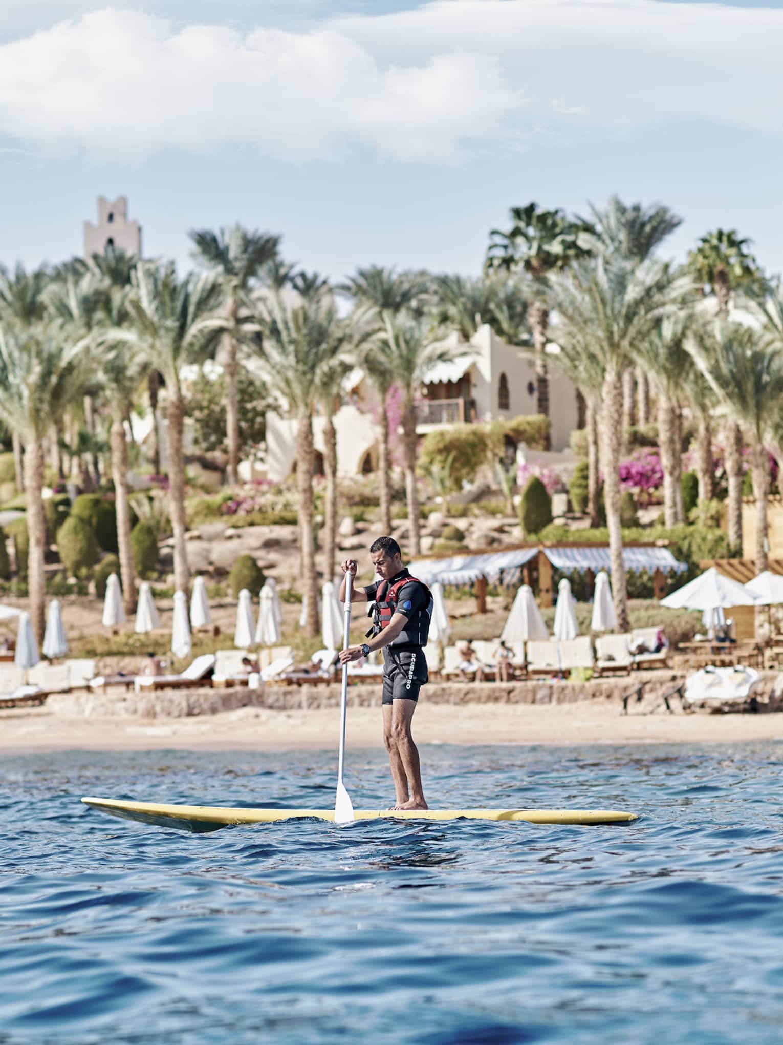 A paddler stands on a board in the water in front of a beach lined with lounge chairs and palm trees and a villa beyond.