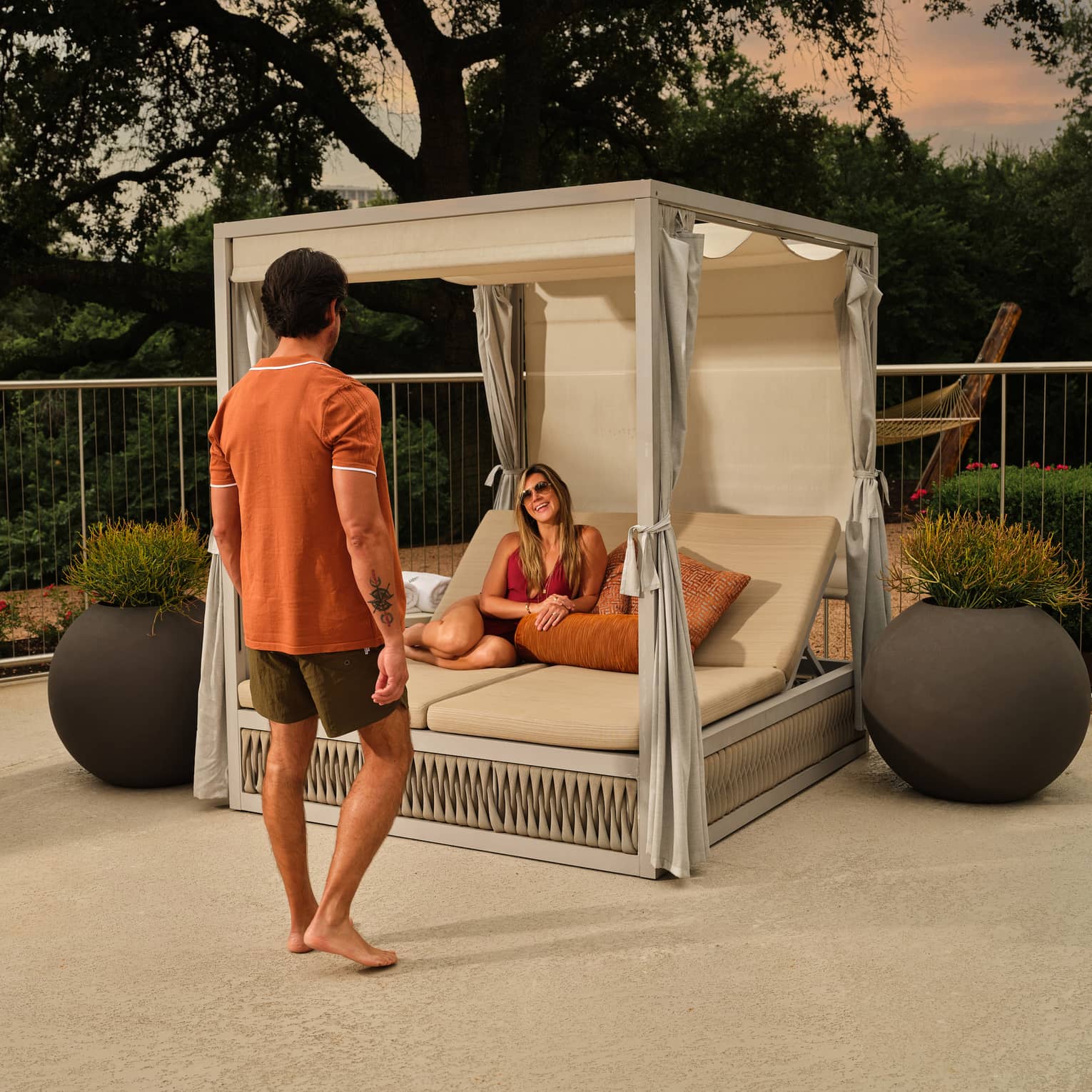 A man walks towards a woman lounging in a cabana surrounded by large potted plants