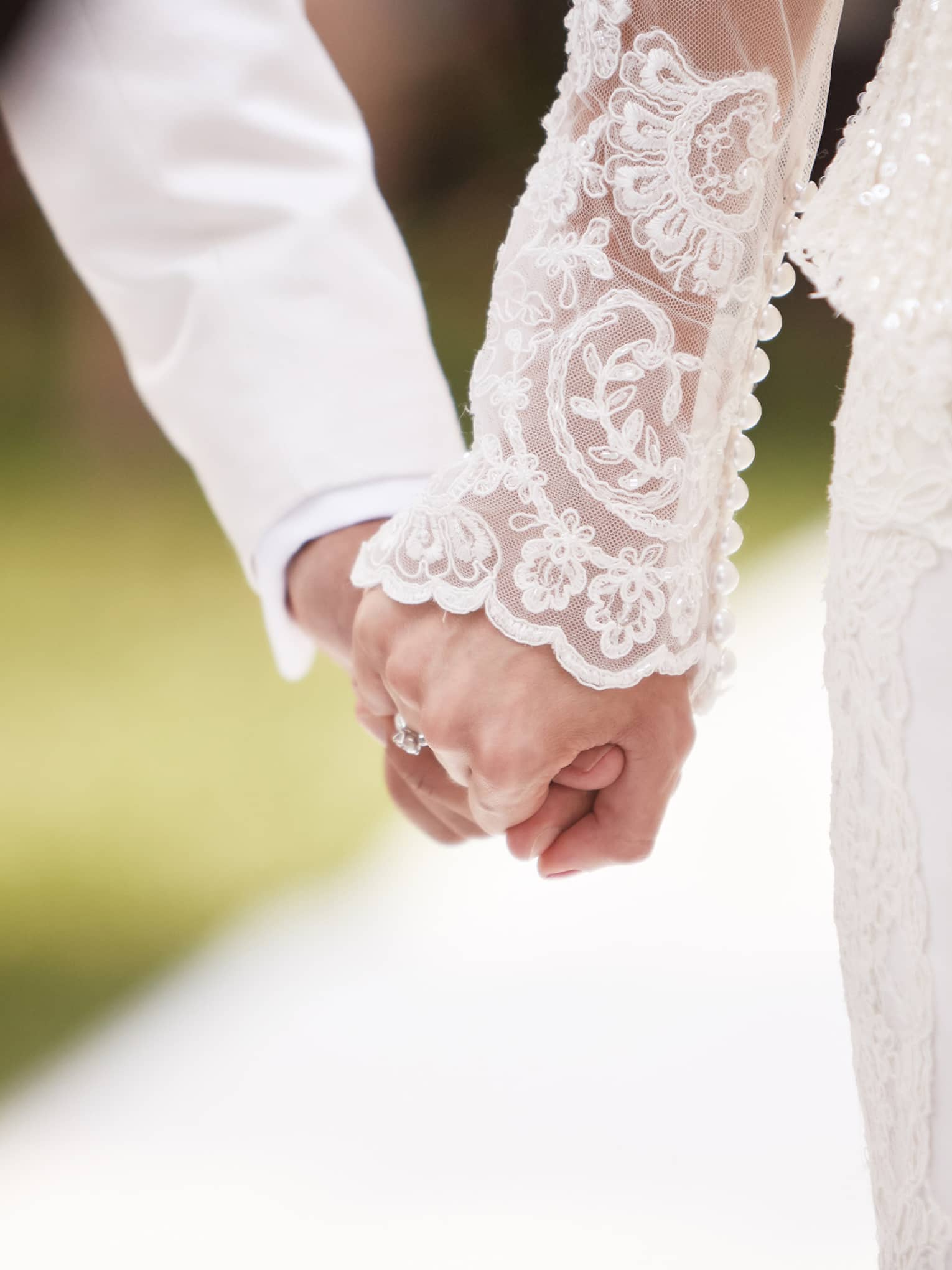 Close-up of bride with lace sleeve and groom with white suit holding hands 