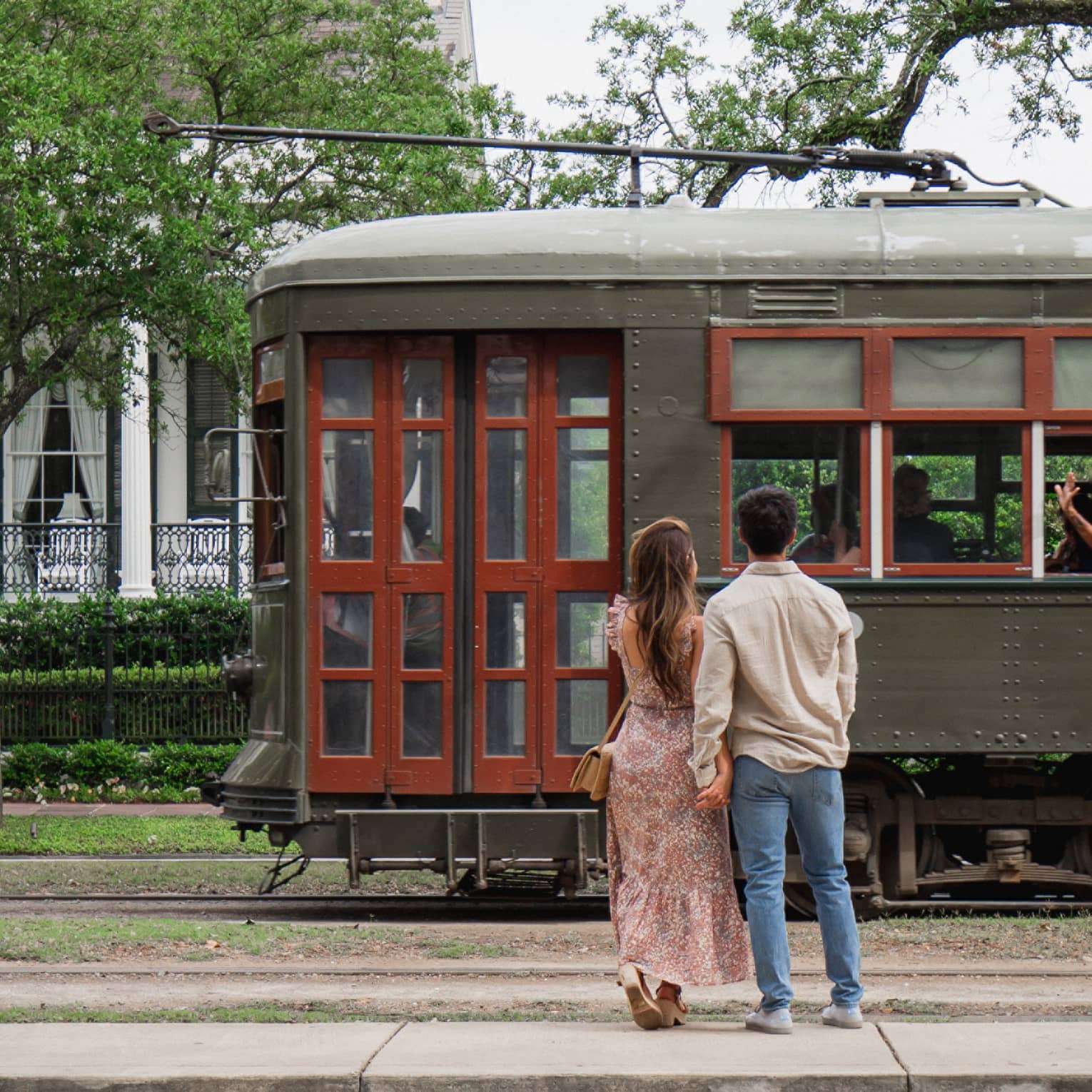 Woman and man hold hands in front of old-fashioned streetcar on New Orleans street, white house in background