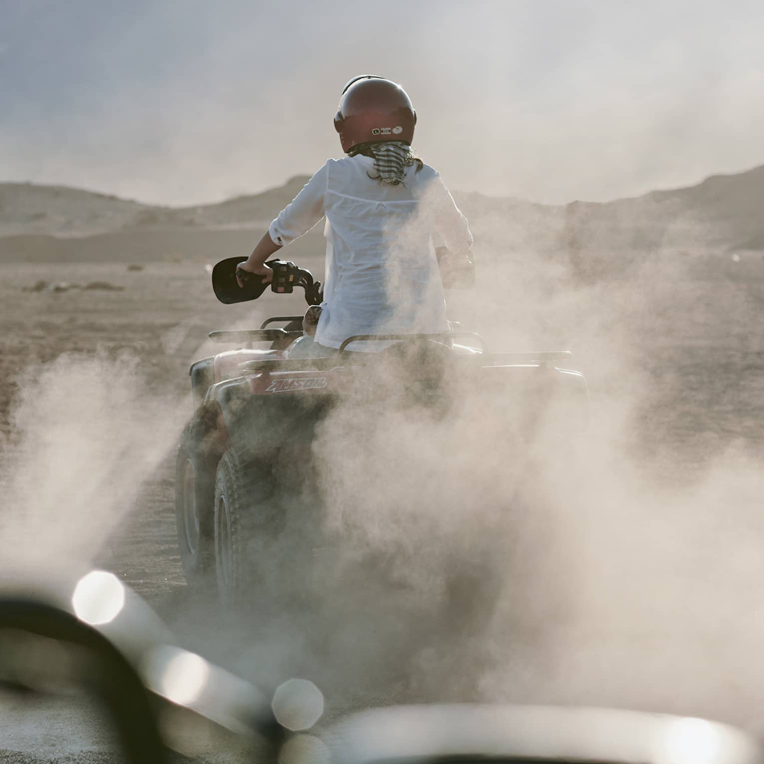Rear view of an adventurer donning a red helmet while riding an ATV amid dust clouds; desert mountains ahead in the distance.